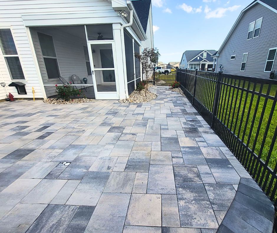 Square rectangle paver stone patio with an aluminum fence to the right and a white house with river rocks in front of it in the background