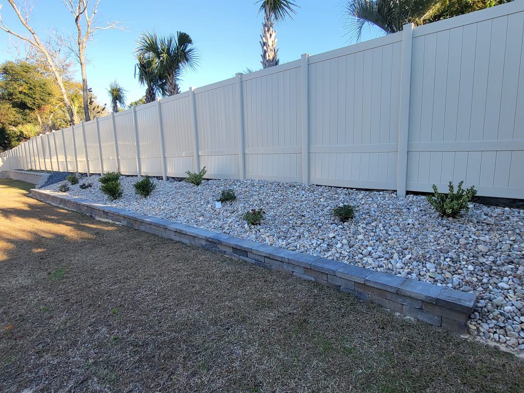 White vinyl privacy fence with white river rock and plants. You can see palm trees behind the fence
