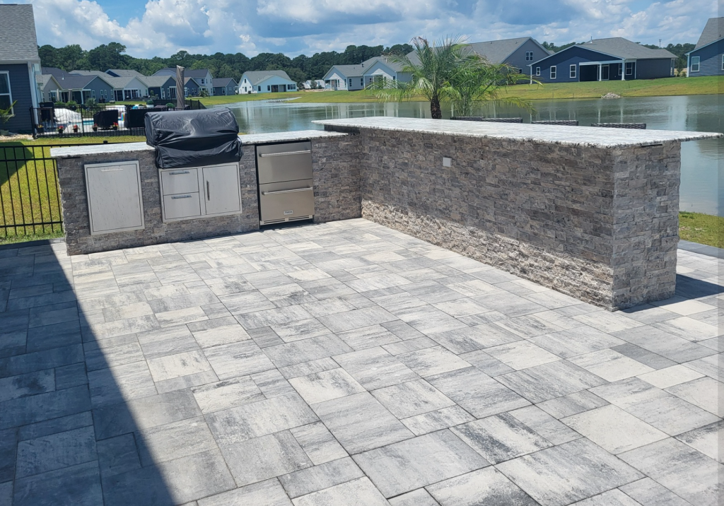 Newly installed or freshly sealed gray paver patio and outdoor kitchen by a scenic lake, demonstrating a clean and protected outdoor living space by Elite Coastal Landscpaing
